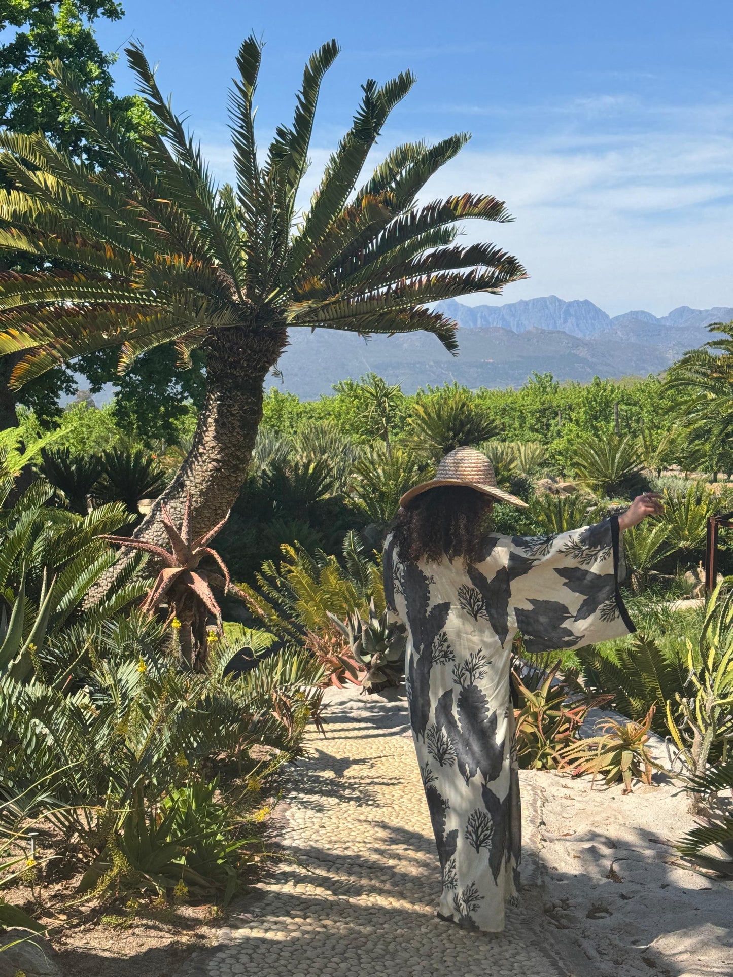 Person in a patterned kimono and hat standing in a garden with palm trees and mountains in the background
