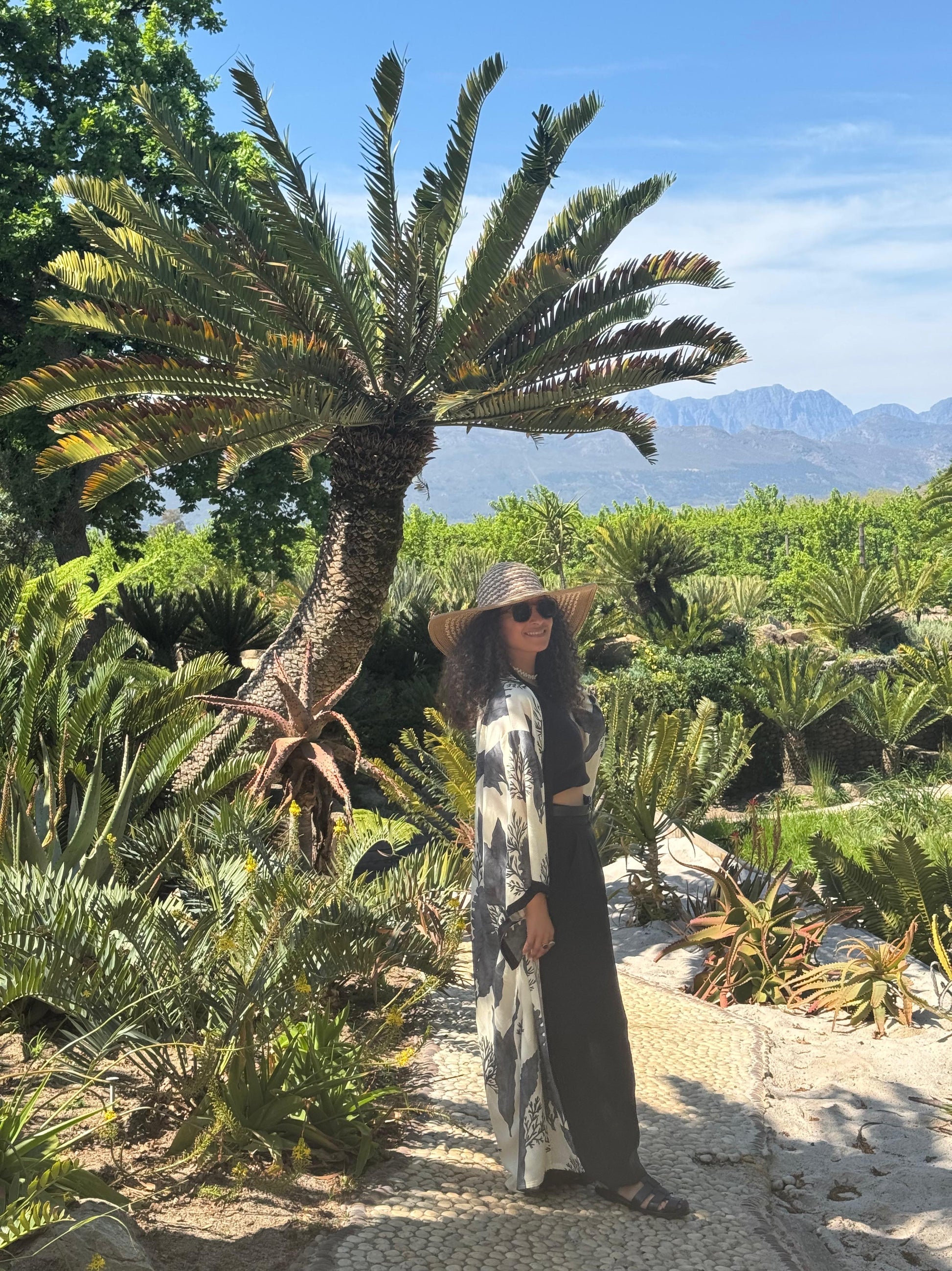 Person standing in a garden with palm trees and mountains in the background